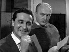 Steed in foreground left chuckles as he chats to Harvey, behind and to his left and frowning