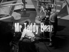 title card: white text reading 'Mr Teddy Bear' superimposed on a wide shot of a TV studio, the crew crouched around the fallen Wayne-Gilley
