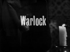 title card: white text reading 'Warlock' superimposed on a dark close-up of the cowled Gallion, his face partly lit by a candle held in his hand