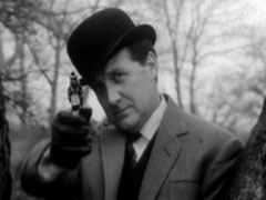 Steed, leaning on the fork of a tree, aims his revolver; he’s dressed in a check blazer and black bowler hat