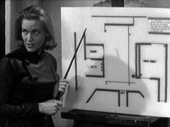Cathy, on the left, wearing her leather flying suit, explains her heist plan to the gang, using a floor plan of the vault