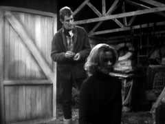 Cathy kneels in front of Quentin, facing us, as Quentin prepares to set fire to the hut and her with it
