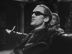 The strange young man flings his arms wide as he regards the interior of the house. He is wearing a leather jacket and sunglasses