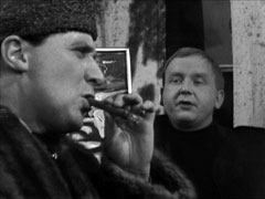 Steed in profile on the left, smoking a cigar as he assumes the guise of an Icelandic art dealer, the artist Leeson faces us from behind him