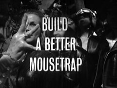 title card: white all caps text reading ‘BUILD A BETTER MOUSETRAP’ superimposed on the biker gang laughing as Ermyntrude tries to put a hex on them
