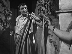 Steed, wearing a toga, is held at gunpoint by Apollodorus