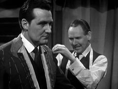 Steed in the foreground wearing a pinned and chalk-marked jacket as Lovell measures him for a suit