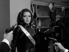 Mrs Peel backs against a pillar as the villains approach her with their guns pointed at her