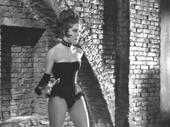 Mrs. Peel, wearing only a black corset and underwear, swings the chain attached to the collar around her neck as she fights Pierre in the catacombs