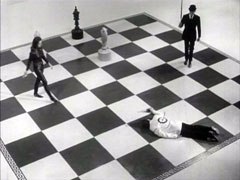 View of an outsized chessboard, skewed from the white queen’s square. The dead man lies across the black diagonal across the king and queen square for the white side while Emma and Steed approach it along the bishop and queen white diagonals; the black king stands on his square, there is a white knight at WK6 and bishop at WQB7
