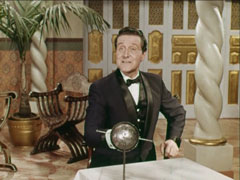 Steed sits at a table in the ornate room, wearing a dinner suit and holding a fencing rapier
