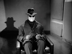 The inert cybernaut sits in an armchair, dressed in a suit, coat, trilby hat, gloves and sunglasses. The room behind is bare and utilitarian, he casts a dramatic shadow toward the left