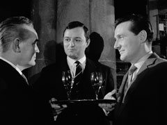 The wine duel begins in the cellar: Harvey faces us holding a tray with two glasses of red wine, he smiles at Boardman to his right. Boardman and Steed are both in profile, facing each other, Boardman on the left
