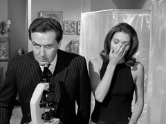 Steed, in his dashing chalk-stripe suit, looks concerned about Emma who is yawning loudly - she wears a tight sleeveless top and stands before the plastic screen shielding the sleeping patients