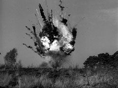 The explosion of the bomb can be seen above the grass at to top of a small hill with trees in the background