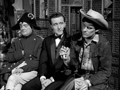 Steed, wearing a tuxedo, sits on a bench between men dressed as Napoleon and a cowboy. A levantine wooden screen stands behind them