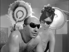 Arkadi lies on a tanning bed as his shapely masseuse attends to him, they are both wearing sunglasses and she fans him with an ostrich feather fan