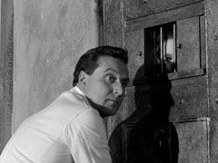 Steed turns to look towards us - and the oncoming guards, as he is unlocking Emma’s cell door; she peers through the grilled window