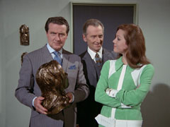 Steed holds the copper bust of Emma as he stands next to her in Beresford’s study, she is smiling at him because Beresford has just made the same quip about preferring the original that he just made