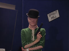 Emma stands in the blue warehouse wearing a green catsuit with lace-up sleeves, her arms are crossed and she holds a pistol in her right hand, pointed over over her left shoulder at a sign that reads SPOUT HILL. A stocking is over her head and she tops her outfit with a black bowler at a jaunty angle
