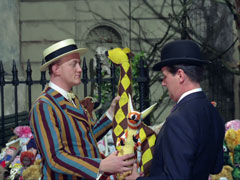 Steed, on the right, smiles at the stuffed kangaroo toy striped in orange and peach that he has just been given by Parker, who wears a striped blazer and boater