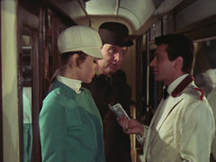 The attendant hands Emma a £5 note in the carriage corridor as Steed looks on