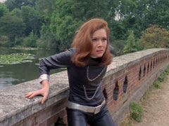 Emma backs against the bricks of a bridge over a lake, she wears a shiny black leather catsuit with silver trim and two chains across the chest