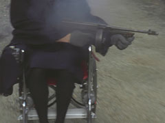 Close-up of the old lady in the wheelchair brandishing a tommy gun - her face is not in frame so you can’t tell who she is