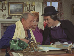 Goat, disguised as Nanny Roberts, hands General Wilmot a pistol to kill Steed after he’s shown the location of the missile bases on a map; Miss Lister sits between them