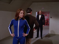 Emma and Steed stand back to back with hands on hips, he facing away, as Professor Stone approaches from behind; Penrose is off-screen approaching from behind the camera.