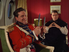 Steed, seated in the aeroplane and dressed as one of Wellington’s officers, straightens a bent poker as Dayton looks on thoughtfully