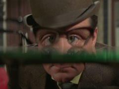 Steed peers to look at some of the spectacles on a shelf, his eye distorted and enlarged as we see them through the lenses
