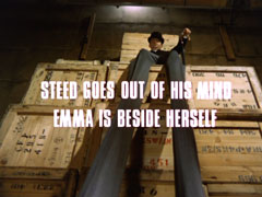 subtitle card: white all caps text reading ‘STEED GOES OUT OF HIS MIND
			EMMA IS BESIDE HERSELF’ superimposed on a low angle view of the enormously long stilt legs of the dead Hooper, perched up on a stack of crates in a warehouse