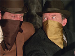 Steed and Merlin smile at each other after defeating the Brigadier with the sleeping gas, handkerchiefs tied around their faces to protect against it