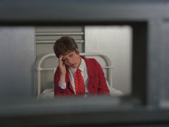 A view through the narrow window of a reinforced cell door - Tara in a red suit & tie holds her head as she sits on the bed