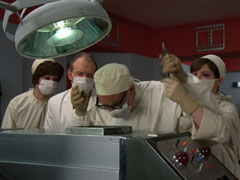 Ardmore ‘operates’ on George, whose quasi-anthropomorhpic casing lies on a surgical gurney under an operating lamp, Tobin stands behind him and they are flanked by the two nurses; all are wearing white surgical gowns, caps and masks