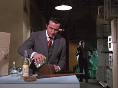 Steed pours the invisibility liquid from the bottom half of the trick Vodka bottle over his bowler hat as Magnus tries to sneak up on him from behind