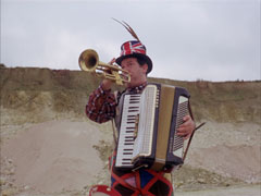 Izzy Pound marches around the quarry practicing his one-man band act. He wears a Union Jack hat with a pheasant feather in it and plays the trumpet and accordion