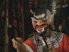 The demon-masked and red robed Ho Lung attacks Tara, a green Chinese tapestry hangs behind him