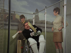 Mother is in the nets, practicing cricket in his wheelchair. He wears pads and gloves and wields a bat. Steed and Tara look on from behind the nets, the wall behind them painted to resemble the view from a village cricket oval with a pavilion and a church tower in the distance