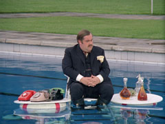 Mother’s office is in a swimming pool - he sits, wearing a suit and tie, on a clear plastic inflatable chair while two floating tables flank him - on the left are four telephones, on the right an assortment of decanters of alcohol and a soda siphon