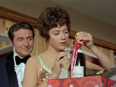 Tara examines the gemstone attached by a ribbon to a bottle of champagne - a gift from Steed, who stands behind her