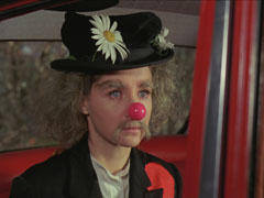 Miranda sits in the back of Steed’s car in her clown disguise; he’s just suggested she remove it.