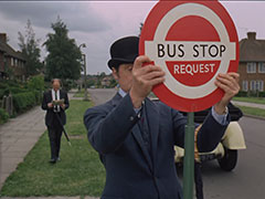 In a quiet suburban street, Steed sets up his own personal bus stop. His yellow Rolls-Royce and a soon-to-be-confused commuter in the background
